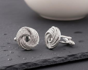 May include: A pair of silver-toned cufflinks featuring a circular design with a textured, rope-like pattern. Each cufflink has a polished metallic finish and a standard toggle closure mechanism on the back, displaying a detailed, twisted knot motif.