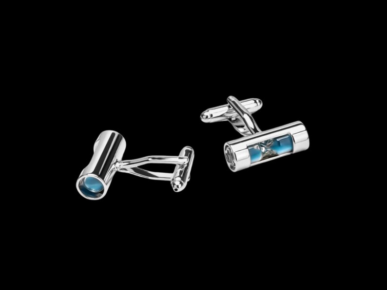 May include: A pair of silver-toned metal cufflinks featuring a cylindrical design. Each cufflink contains a small, clear glass tube filled with blue sand, resembling a miniature hourglass, with a standard hinged toggle closure on the back.