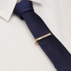 May include: A navy blue tie with a gold tie bar featuring a rectangular mother-of-pearl inlay. The tie is paired with a white dress shirt, creating a classic and elegant look.