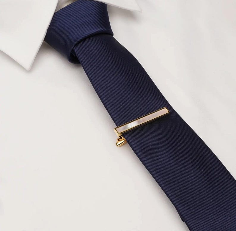 May include: A navy blue tie with a gold tie bar featuring a rectangular mother-of-pearl inlay. The tie is paired with a white dress shirt, creating a classic and elegant look.
