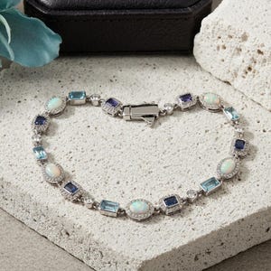 May include: A silver-toned bracelet featuring alternating oval-cut opals, rectangular-cut blue topaz, and rectangular-cut dark blue sapphires. Each gemstone is surrounded by small clear stones, connected by silver links with a secure metal clasp.