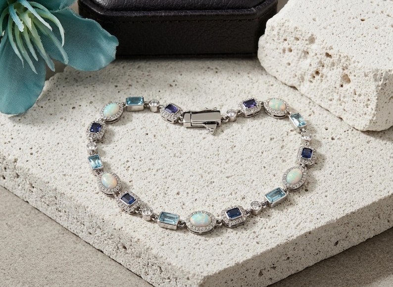 May include: A silver-toned bracelet featuring alternating oval-cut opals, rectangular-cut blue topaz, and rectangular-cut dark blue sapphires. Each gemstone is surrounded by small clear stones, connected by silver links with a secure metal clasp.