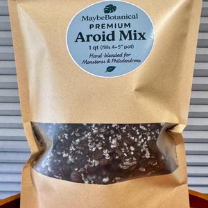 May include: A brown paper bag with a clear window, filled with a dark soil mix. A light blue circular label reads "MaybeBotanical PREMIUM Aroid Mix" and specifies the contents are hand-blended for Monsteras & Philodendrons, filling a 4-5 inch pot.