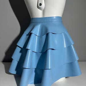 May include: A light blue latex skirt featuring a high waistband and three distinct horizontal tiers of ruffled fabric. The garment has a smooth, semi-glossy finish and a flared silhouette that extends from the waist down to the hemline.