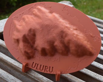 May include: A reddish-brown, oval-shaped 3D topographical map of Uluru, Australia, with the word "ULURU" at the base. The map is mounted on a small stand and sits on a wooden surface.