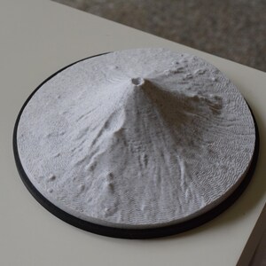 May include: A white, circular relief sculpture of a mountain with a central crater. The sculpture has a textured surface and sits on a black circular base. It is displayed on a light-coloured surface.