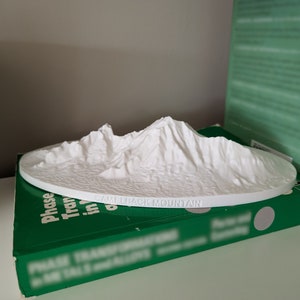 May include: A white 3D-printed model of Camelback Mountain. The model is on a white circular base, with the mountain's peaks and terrain detailed. The text "Camelback Mountain" is printed on the base. The model is resting on a green book.