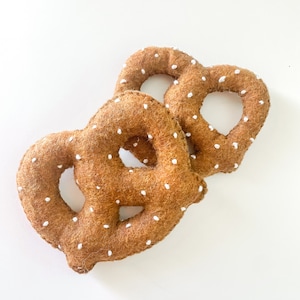 May include: Two soft pretzels with a brown felt texture and white dotted details. The pretzels are in a heart shape, and are placed on a white background. The image is a close-up shot of the pretzels.