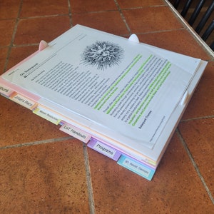 Puede incluir: Una pila de papeles con pestañas de colores etiquetadas como "Solarpunk", "Jillian's Recs", "Maker Resources", "LOT Handouts", "Programs" y "30 Assist Devices". El papel superior tiene una ilustración en blanco y negro de un sol con una cara y el texto "Solarpunk Themes".