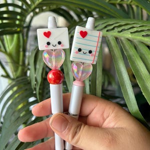 May include: Two white pens with cute, colorful, clay charms attached to the top. The charms are shaped like envelopes and a piece of paper with red hearts and smiley faces. The pens are held in a hand.