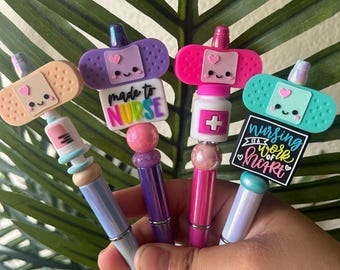 May include: A collection of decorative pens with medical-themed toppers. The pens feature designs like bandages, syringes, and pill bottles, with the phrases "made to nurse" and "nursing is a work of heart". The pens have various colors, including pink, purple, and blue.