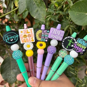 May include: Six colorful pens with different inspirational messages and charms. The pens are decorated with rhinestones, flowers, and other embellishments. The messages on the pens include "Stronger than the storm", "Only good vibes", "She believed she could so she did", "Kindness matters", "It makes them positive", and "Dare to dream".