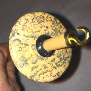 Könnte beinhalten: Ein Spinnrad-Whorl mit einem gesprenkelten, gelben und schwarzen Design. Der Whorl ist aus Holz und hat eine schwarze und goldene Metallspindel.