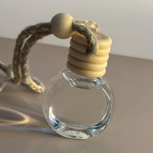 Puede incluir: Una botella de vidrio transparente y redonda con una tapa de madera y una cuerda beige para colgar. La botella está llena de un líquido transparente. La tapa de madera tiene un diseño acanalado. La cuerda tiene detalles dorados.