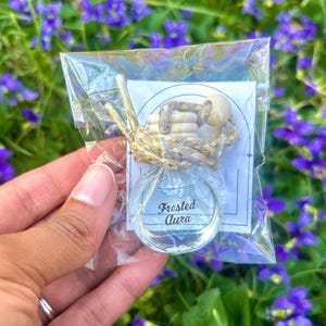 May include: A clear glass bottle with the text "Frosted Aura" is packaged in a clear plastic bag. The bottle is attached to a decorative wooden and rope accent. The background features green grass and purple flowers.