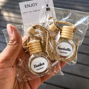 May include: Two small, clear glass car air fresheners in plastic packaging. Each has a wooden cap and a decorative rope. One label reads "Vanber custom," the other "Sandnilla custom." A card reads "ENJOY your car diffuser."