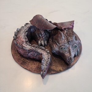 May include: A detailed sculpture of a curled-up dragon with a textured, cracked surface. The dragon is primarily bronze and silver with hints of purple. It has green eyes and is resting on a bronze-colored circular base. The sculpture is a fantasy-themed decorative item.