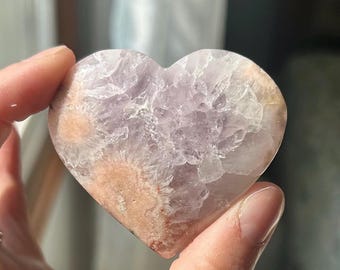 May include: A heart-shaped crystal stone with a mix of lavender, white, and peach colors. The stone has a textured surface and is held in a hand. The crystal is a decorative item.