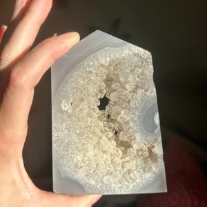 May include: A white agate geode with a large opening revealing a cluster of small, clear quartz crystals. The geode is shaped like a house and has a smooth, polished surface.