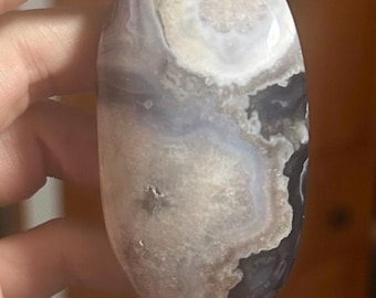 May include: An oval-shaped polished stone with a mix of colors. The stone features a blend of white, gray, and black hues, with a marbled appearance. The stone is held between fingers, showcasing its smooth, rounded form. The stone's surface has a glossy finish.