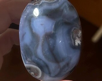 May include: A polished, oval-shaped stone with a swirling pattern of blue, gray, and white. The stone has a smooth, glossy surface and is held in a hand. The stone's colors and patterns create a unique, natural aesthetic.