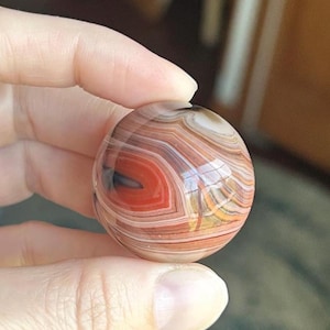 May include: A polished, spherical agate stone with bands of red, brown, white, and black. The gemstone is held between fingers, showcasing its smooth, reflective surface. The agate's natural patterns create a unique, swirling design.