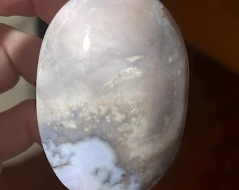 May include: An oval-shaped, polished stone with a blend of soft lavender, gray, and white hues. The stone features a cloudy, layered pattern with areas of subtle banding and a smooth, glossy surface. The overall aesthetic is natural and organic.