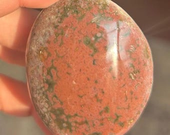 Può includere: Una pietra levigata di forma ovale con un motivo screziato. La pietra presenta un colore di base marrone-rossastro con venature verdi e grigie. La superficie è liscia e riflettente, indicando che è stata lucidata.