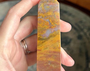 May include: A rectangular, polished stone obelisk with a pointed top. The stone features a blend of warm colors, including shades of yellow, orange, and brown, with streaks of gray and purple. The obelisk is held in a hand.
