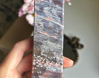 Puede incluir: Un obelisco de piedra rectangular y pulida con una parte superior puntiaguda. La piedra presenta una mezcla de colores, incluyendo rojo intenso, gris, blanco y toques de naranja. La superficie tiene un aspecto veteado y estratificado, con pequeños patrones moteados.