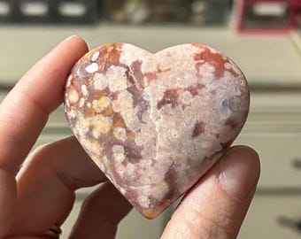 May include: A heart-shaped pink and orange stone with white and brown markings. The stone is polished and smooth.
