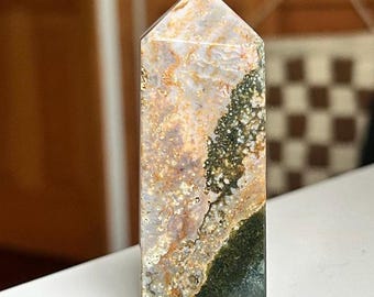 May include: A tall, rectangular crystal obelisk with a pointed top. The stone features a mix of colors, including beige, brown, green, and white, with a marbled appearance. The obelisk is standing upright on a white surface.