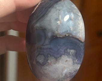 May include: An oval-shaped, polished stone with a mix of blue, gray, and white colors. The stone has a smooth surface with visible patterns and textures. The stone is held between fingers.