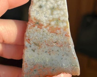 May include: A polished, irregularly shaped stone with a mix of colors. The top portion is white with small, speckled patterns. The middle section has orange and red hues, transitioning to a gray and red base. The stone is held by a hand.