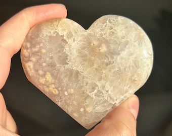 May include: A heart-shaped crystal stone with a light beige and white color scheme. The stone has a textured surface with small, light brown spots. The crystal is held in a hand against a dark background.