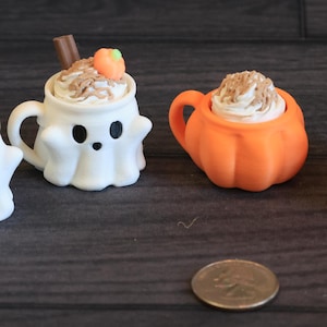 May include: Four miniature Halloween-themed mugs. Two are white ghost-shaped mugs, and two are orange pumpkin-shaped mugs. Each mug is topped with whipped cream and a cinnamon stick. A small coin is in the foreground.