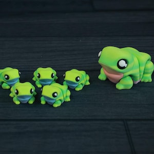 May include: A group of seven green and yellow frog figurines. The two largest frogs are facing each other, while the five smaller frogs are arranged in a semi-circle in front of them.