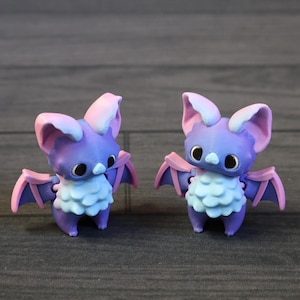 May include: Two purple bat figurines with pink and blue details. The bats have large ears, wings, and a fluffy chest. They are on a grey wooden surface.