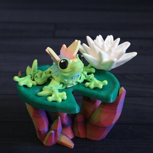 May include: A green frog with rainbow wings sits on a green lily pad with a white water lily. The lily pad and flower are on a colorful, geometric base.