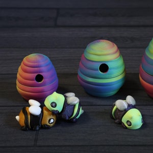 May include: A set of three rainbow-colored beehive-shaped containers with four small bee figurines. The bee figurines are green, black, and white.