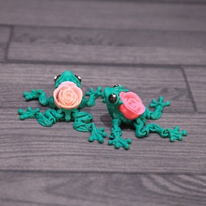 May include: Two green frog figurines with pink roses on their backs. The frogs are posed with their legs outstretched and their heads tilted slightly.