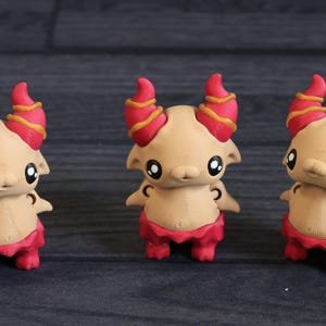 May include: Three tan-colored figurines with red and yellow horns, red skirts, and large black eyes. The figurines have small arms and are standing on a dark surface. The overall theme is fantasy creatures.