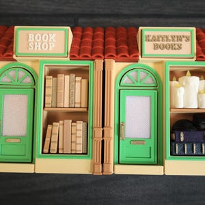 May include: A miniature book shop and bookstore display. The model features two connected buildings with green doors, tan walls, and red tile roofs. One building is labeled "BOOK SHOP" and the other "KAITLYN'S BOOKS". The shop windows display books and candles.