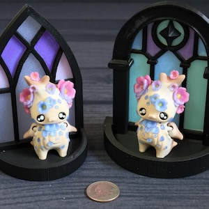 May include: Two whimsical figurines, each with a cream-colored body, blue floral accents, and pink flower crowns, stand in front of black arched window frames with stained glass designs. A coin is at the bottom.