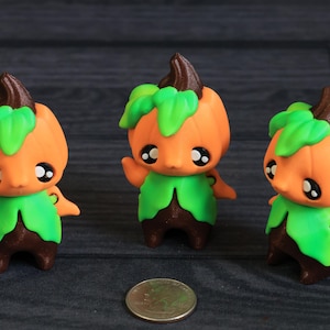 May include: Three orange pumpkin-shaped figurines with green leaf accents and brown stems. Each figure has large, expressive eyes and a green leafy collar. A coin is placed in front of the figurines.