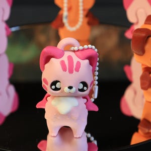 May include: A collection of colorful cat figurines. The central figurine is pink with dark pink accents, large black eyes, and a white chest. Other figurines are orange and pink, with a reflective surface.