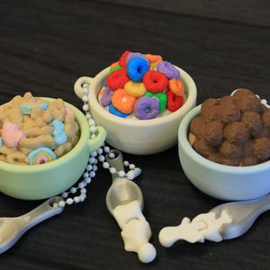 May include: Three miniature cereal bowls with spoons. One bowl contains colorful cereal rings, another has brown cereal balls, and the third has cereal with marshmallows. Each bowl is a different color and has a spoon attached.