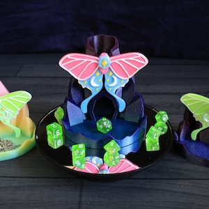 May include: Three decorative tabletop pieces featuring moth designs. The central piece has a pink and blue moth atop a blue structure, surrounded by green dice. The other two pieces have green moths and are in shades of pink, yellow, and blue.