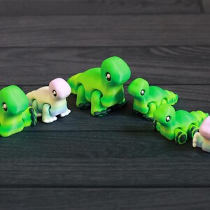 以下が含まれることがあります： 緑とピンクの小さな関節式の恐竜のおもちゃが5つ。恐竜は一列に並んでおり、中央に最も大きな恐竜がいます。