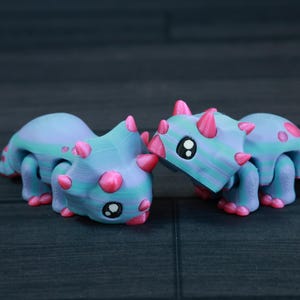 以下が含まれることがあります： 大きな目をした、青とピンクの小さな関節式の恐竜のおもちゃが2つ。恐竜は互いに向き合っており、鼻を触っているように見える。